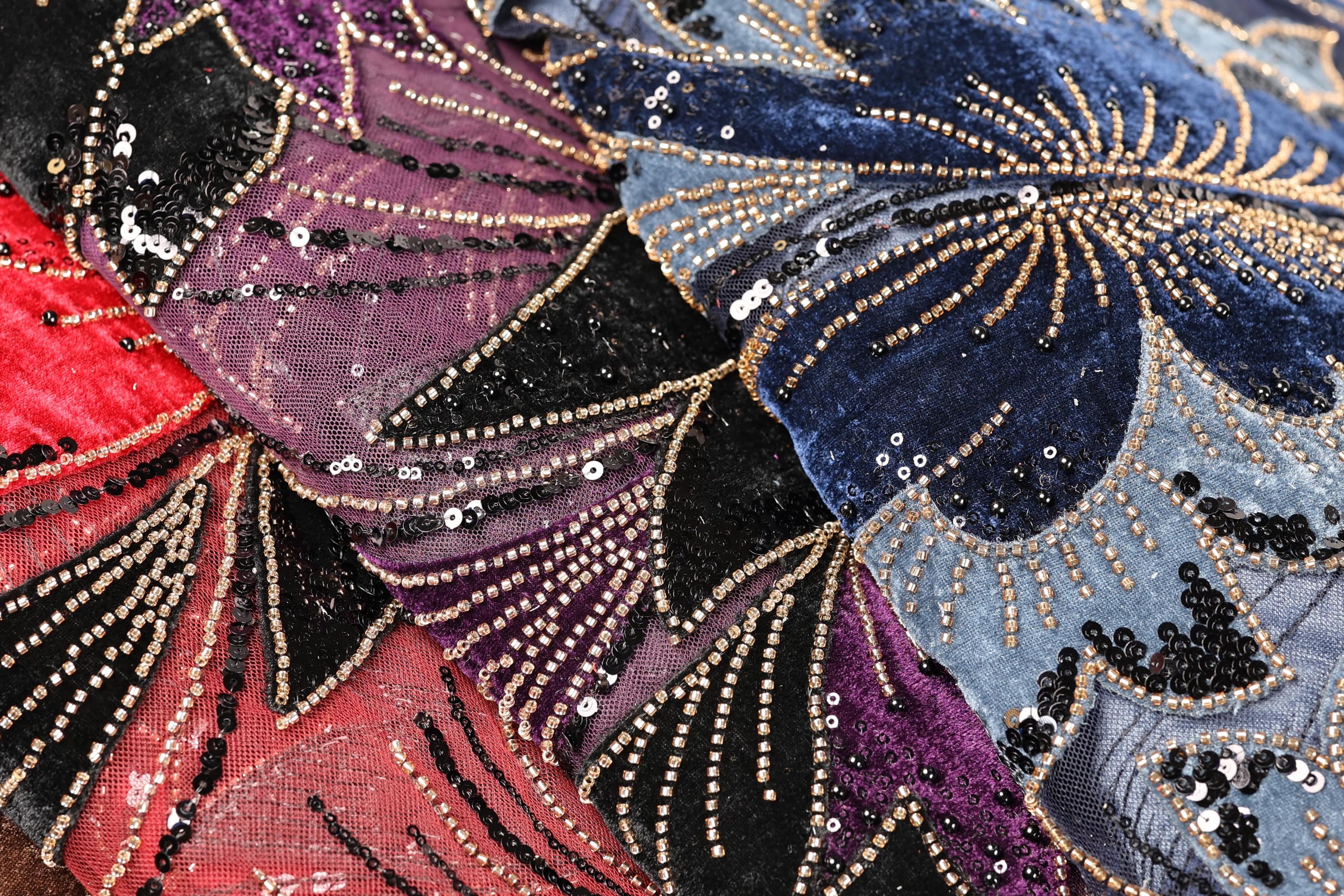 The image is a close-up of a fabric with a vibrant, colorful, and intricate design featuring a variety of sequins and beads in shades of red, blue, and purple.