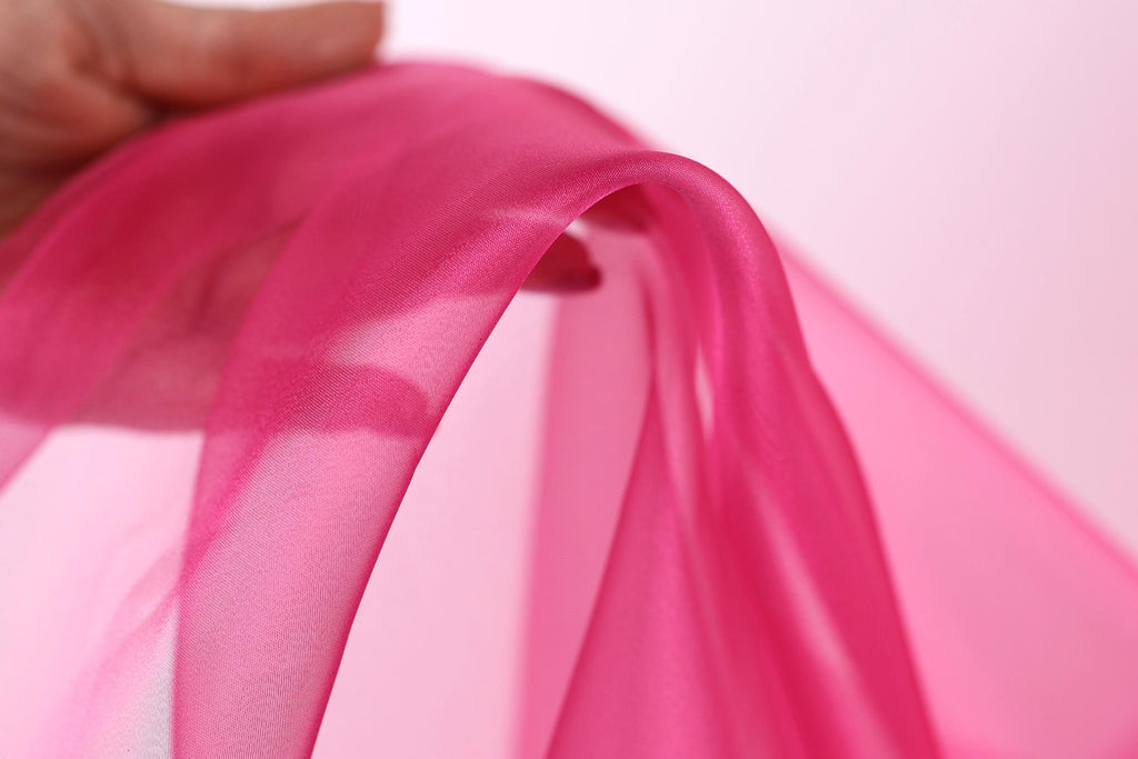 A hand is holding a piece of pink fabric, which appears to be silk or chiffon. The fabric is being held in a curved position, creating a sense of movement and softness.