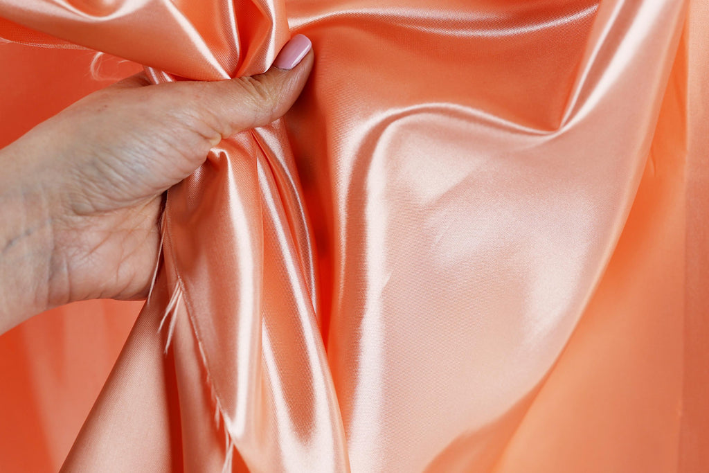 A hand is holding a piece of peach-colored satin fabric, with the fabric being crumpled and wrinkled.