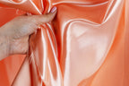 A hand is holding a piece of peach-colored satin fabric, with the fabric being crumpled and wrinkled.