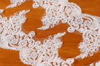 a close-up view of a delicate, intricate white lace fabric with floral and leaf-like patterns. The fabric appears to be a lace trim or a decorative piece, possibly used for a wedding or other special occasions.