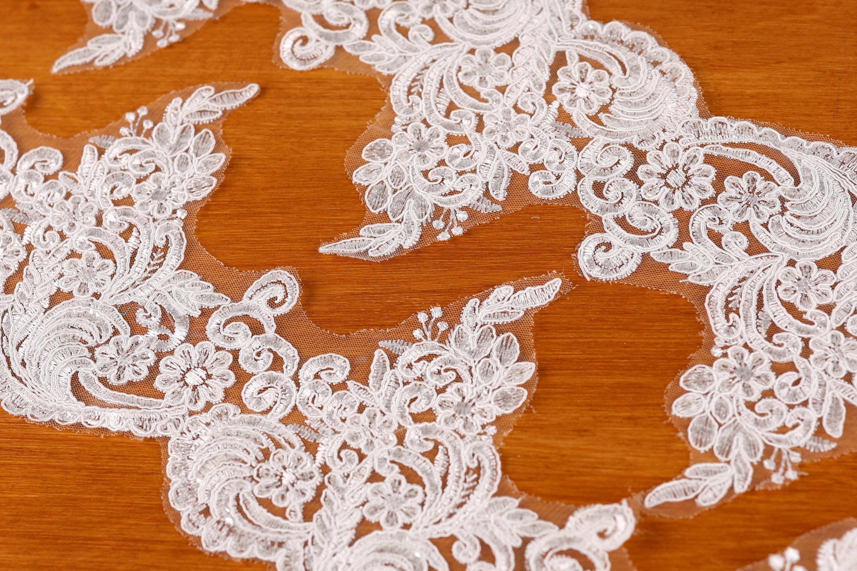 a close-up view of a delicate, intricate white lace fabric with floral and leaf-like patterns. The fabric appears to be a lace trim or a decorative piece, possibly used for a wedding or other special occasions.