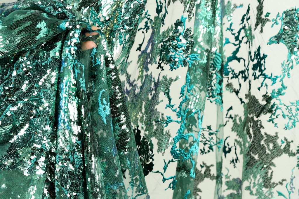 a close-up view of a fabric with a green and blue floral pattern. The fabric appears to be made of sequins or other decorative materials, creating a visually striking and intricate design.
