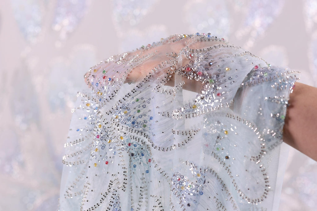 A hand is holding a piece of fabric with a silver and blue floral pattern, and the background is a light blue color with a white floral pattern.
