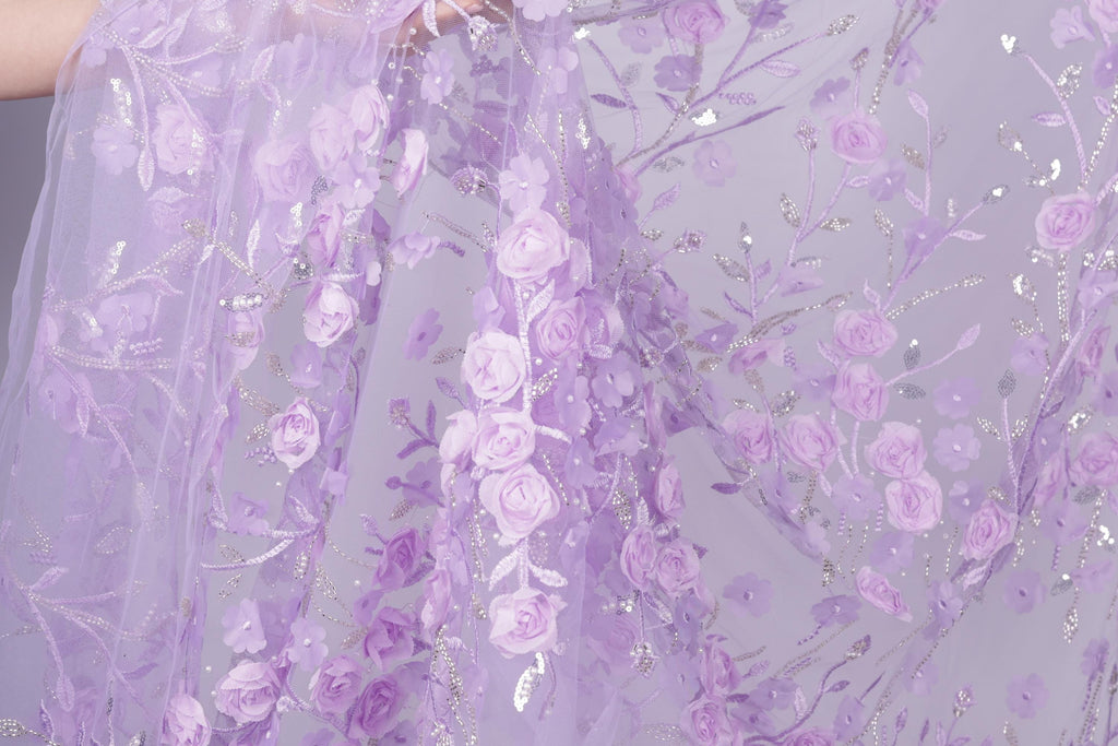a close-up view of a delicate, lavender-colored fabric adorned with a floral pattern of pink roses and leaves. The fabric appears to be made of sheer material, giving it a soft and delicate appearance.