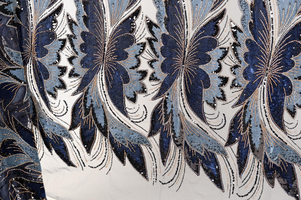 a close-up view of a fabric with a repeating pattern of blue and white butterfly-like shapes, adorned with sequins and beads. The pattern is intricate and detailed, showcasing the craftsmanship and artistry involved in creating the fabric.