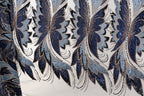 a close-up view of a fabric with a repeating pattern of blue and white butterfly-like shapes, adorned with sequins and beads. The pattern is intricate and detailed, showcasing the craftsmanship and artistry involved in creating the fabric.