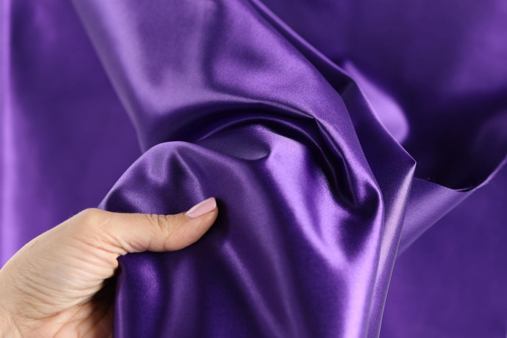 A hand is holding a piece of purple satin fabric, with the fabric's surface wrinkled and shiny.