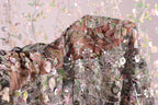 a close-up view of a delicate, intricately embroidered fabric with a floral pattern. The fabric appears to be made of sheer material, allowing the colorful flowers and leaves to be clearly visible. The background is a soft, blurred pink color, which complements the vibrant colors of the embroidered design. The image does not contain any text or other discernible objects, and the focus is solely on the detailed embroidery and the fabric's texture.