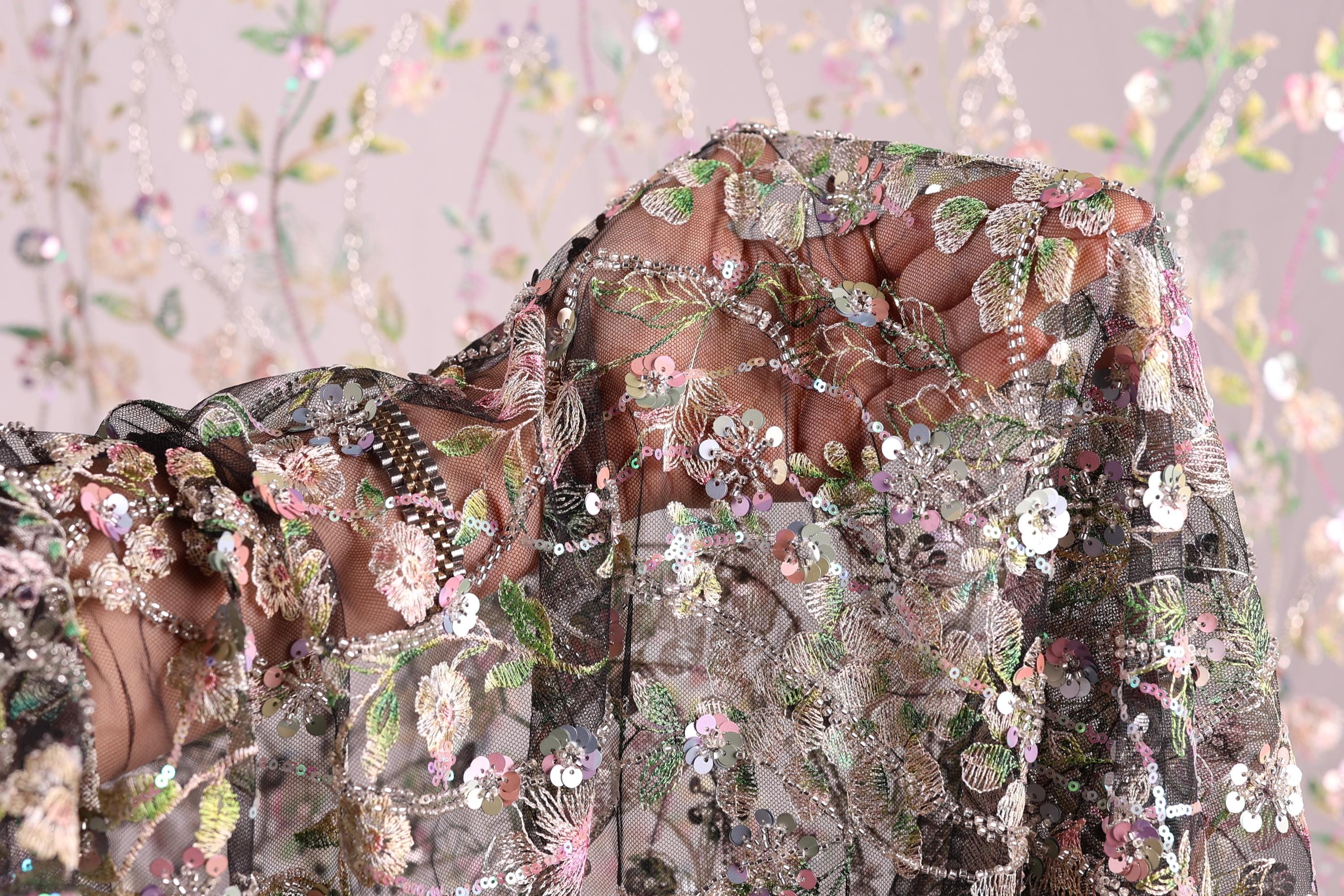a close-up view of a delicate, intricately embroidered fabric with a floral pattern. The fabric appears to be made of sheer material, allowing the colorful flowers and leaves to be clearly visible. The background is a soft, blurred pink color, which complements the vibrant colors of the embroidered design. The image does not contain any text or other discernible objects, and the focus is solely on the detailed embroidery and the fabric's texture.