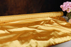 a roll of shiny gold satin fabric, with a vase of pink flowers placed next to it on a dark background.