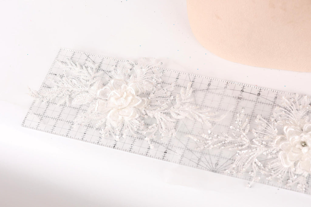 a close-up view of a delicate, white lace flower on a transparent ruler, with a light-colored fabric or material in the background.