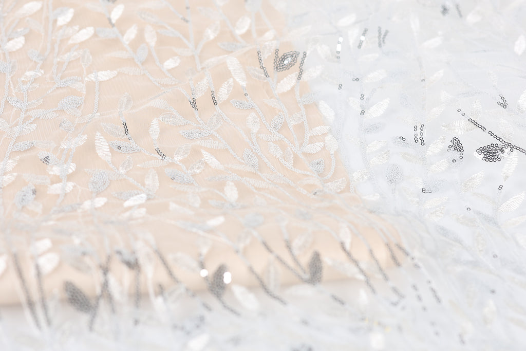 The image is a close-up of a fabric with a floral pattern, featuring a mix of white and beige colors. The fabric appears to be made of lace or a similar delicate material, with intricate white and beige embroidered designs.