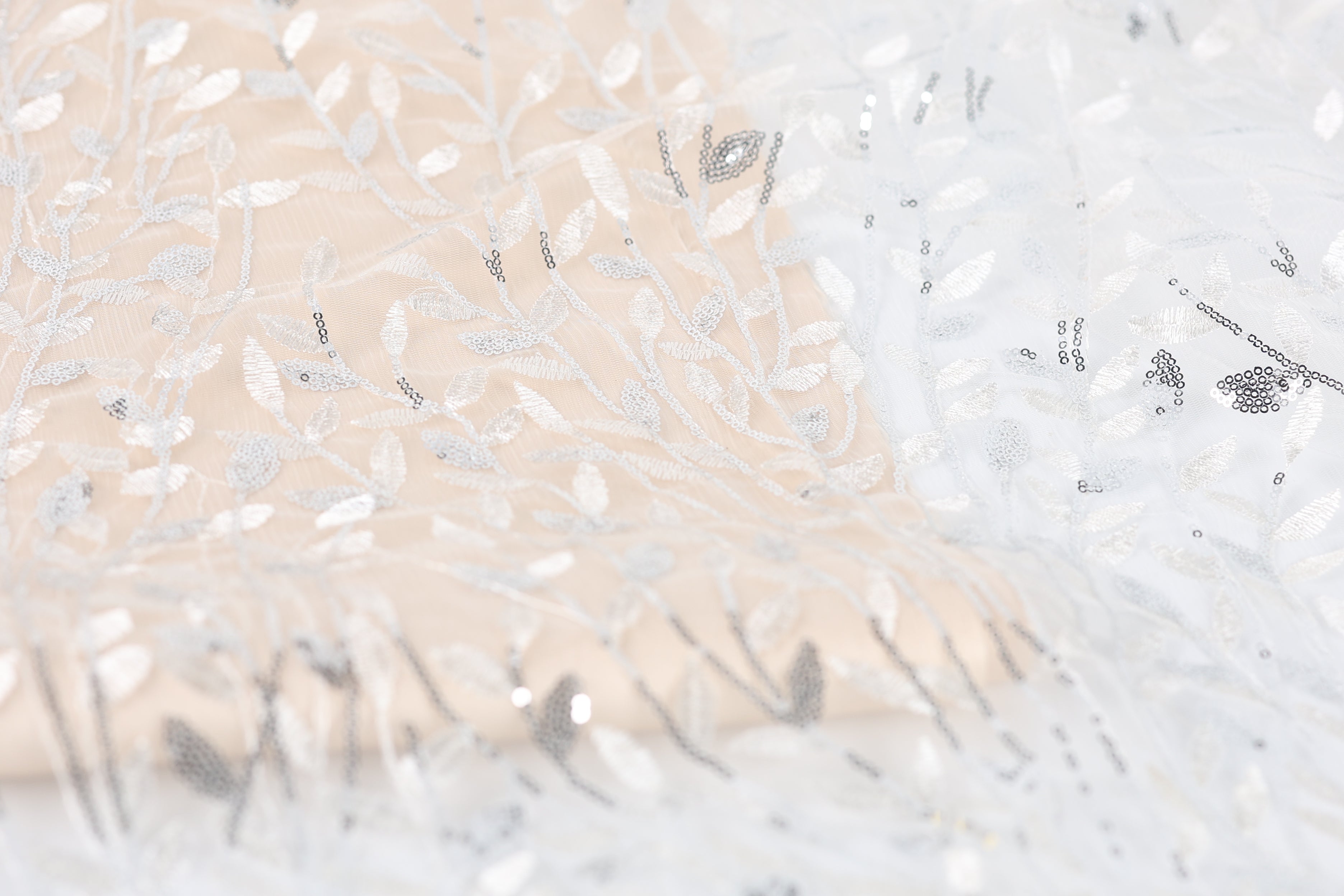 The image is a close-up of a fabric with a floral pattern, featuring a mix of white and beige colors. The fabric appears to be made of lace or a similar delicate material, with intricate white and beige embroidered designs.