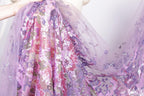 A close-up view of a fabric with a floral pattern in shades of purple and pink, featuring sequins and lace-like details.
