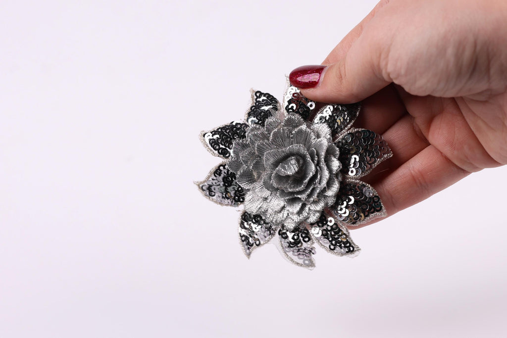 A hand holding a silver flower-shaped ornament with sequins against a white background.