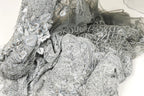 a close-up view of a delicate, intricately designed fabric with a silver and gray color scheme. The fabric appears to be made of lace or a similar material, with a floral or leaf-like pattern visible on the surface. The fabric is crumpled and wrinkled, giving it a textured and somewhat messy appearance.