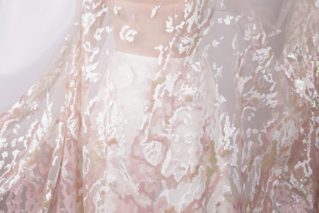 a close-up view of a delicate, lace-like fabric with a floral pattern in shades of pink and white. The fabric appears to be a part of a garment, possibly a dress or a blouse, with a sheer, flowing design. The intricate details and delicate nature of the fabric create a visually appealing and elegant appearance.