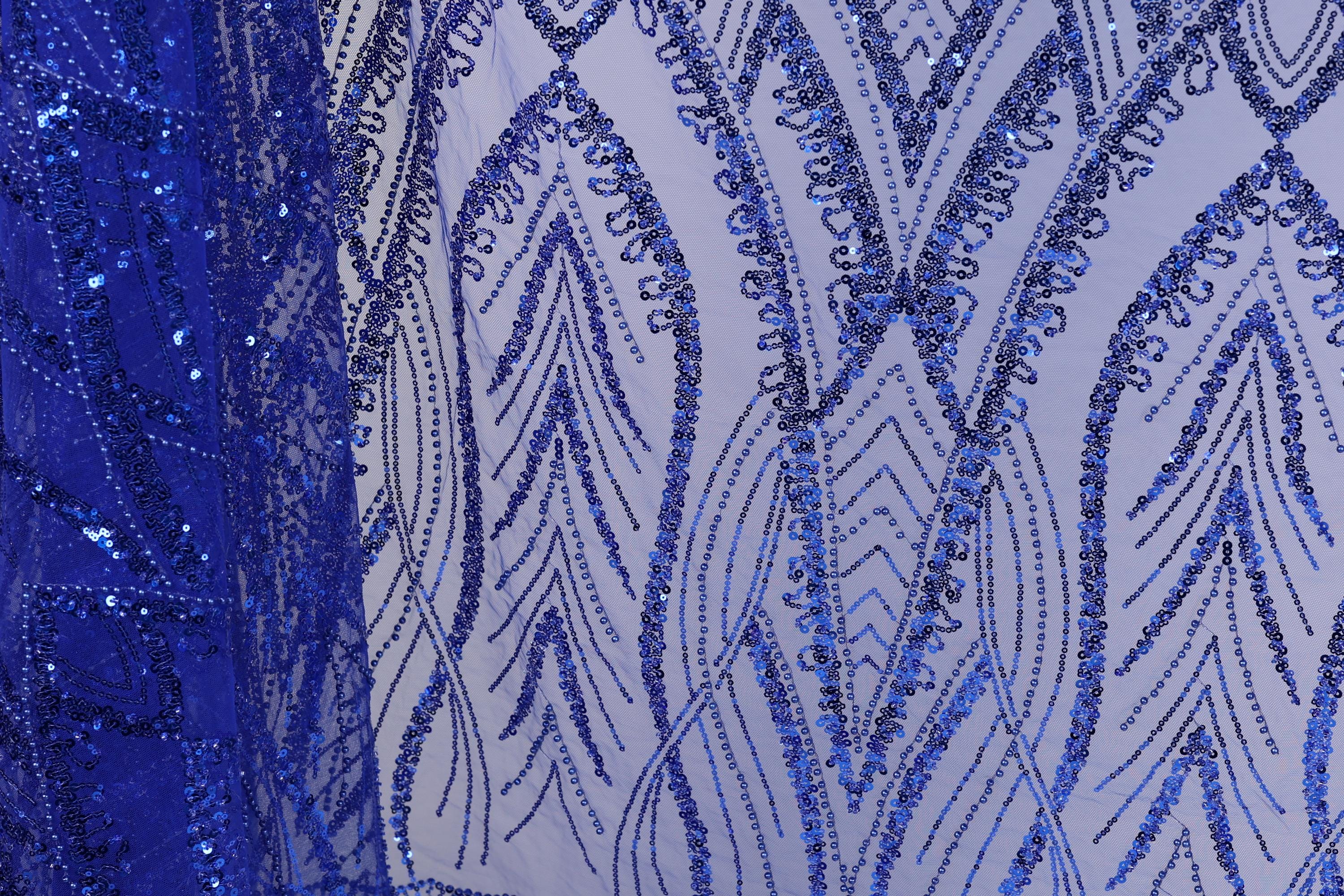 a close-up view of a fabric with a striking blue sequin pattern. The pattern is intricate and detailed, with a repeating design of blue sequins arranged in a zigzag pattern across the fabric. The fabric appears to be a sheer material, allowing the blue sequins to stand out prominently. The image does not contain any text or other discernible objects, and the focus is solely on the detailed blue sequin pattern.