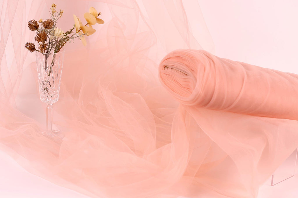 a glass vase with flowers and a rolled-up piece of pink fabric, all set against a soft pink background.