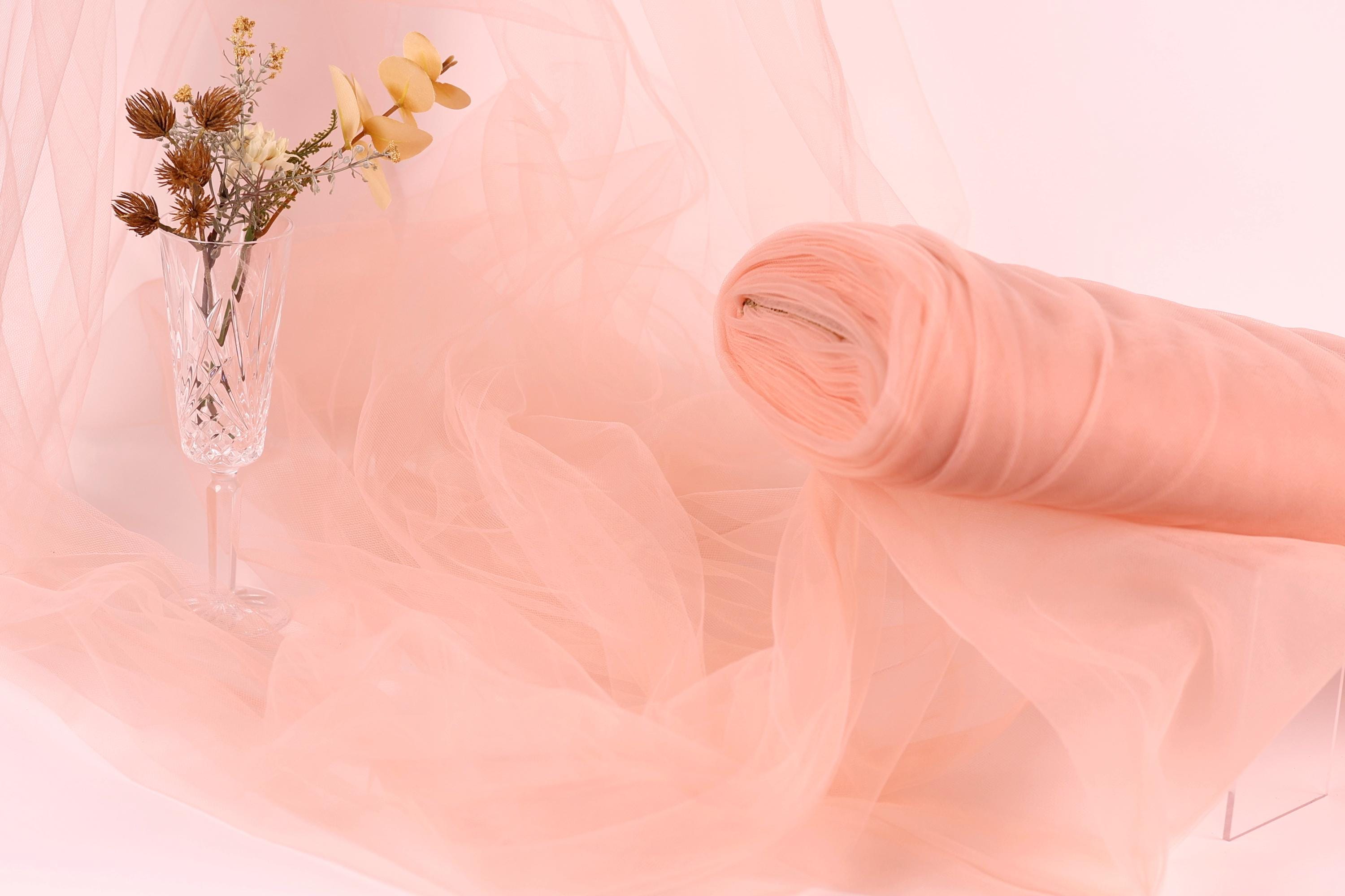 a glass vase with flowers and a rolled-up piece of pink fabric, all set against a soft pink background.