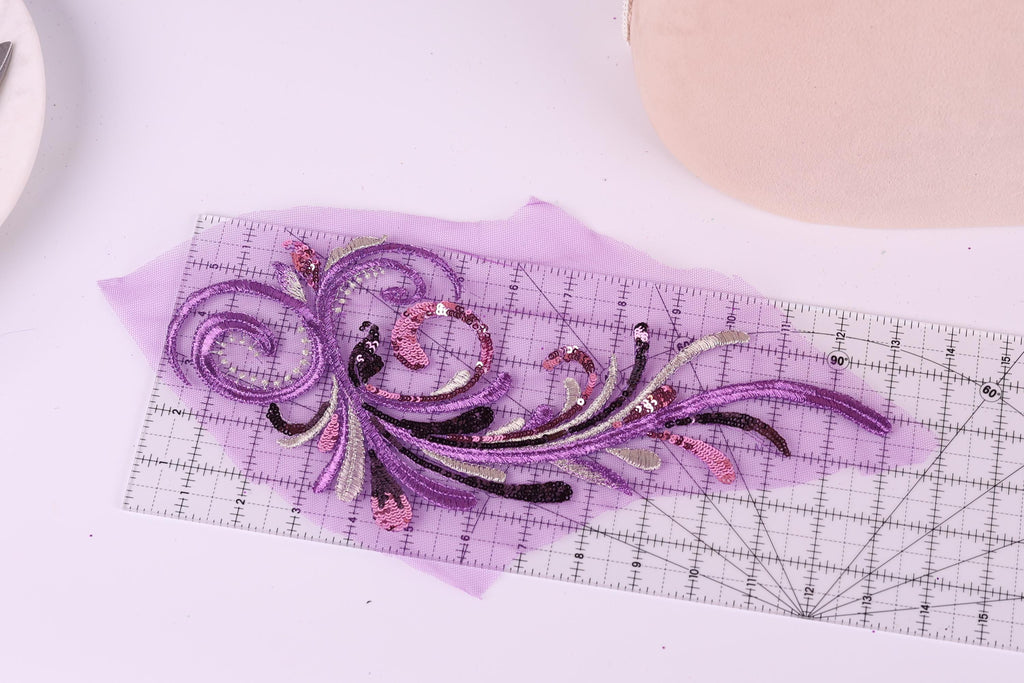a close-up view of a decorative purple and pink embroidered piece on a transparent ruler, with a pink object in the background.