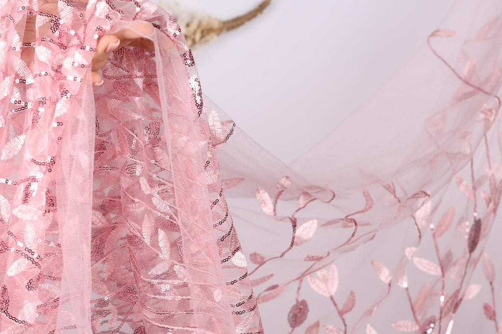 A close-up view of a delicate pink fabric with a floral pattern, featuring a lace-like texture and sequin accents.