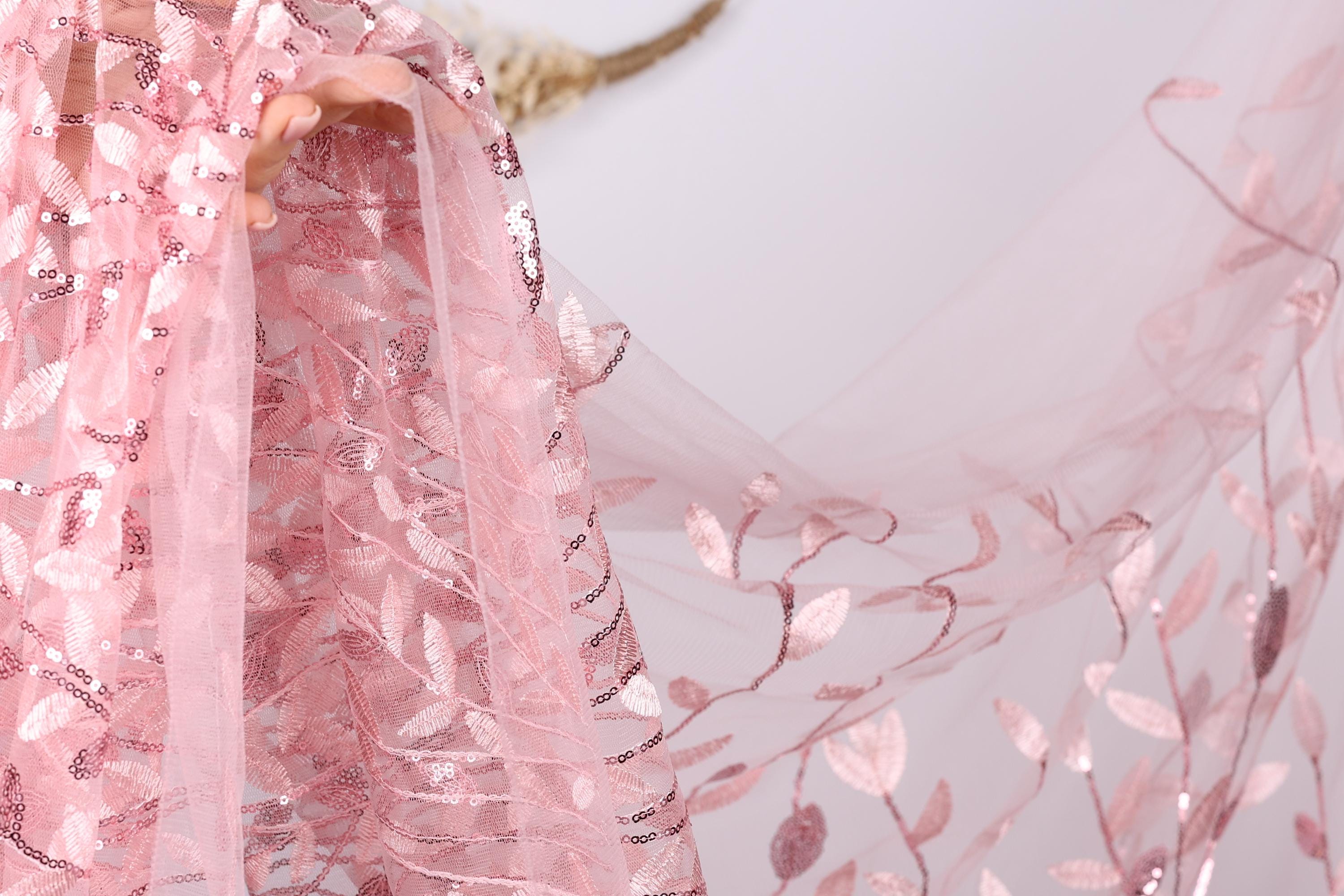 A close-up view of a delicate pink fabric with a floral pattern, featuring a lace-like texture and sequin accents.