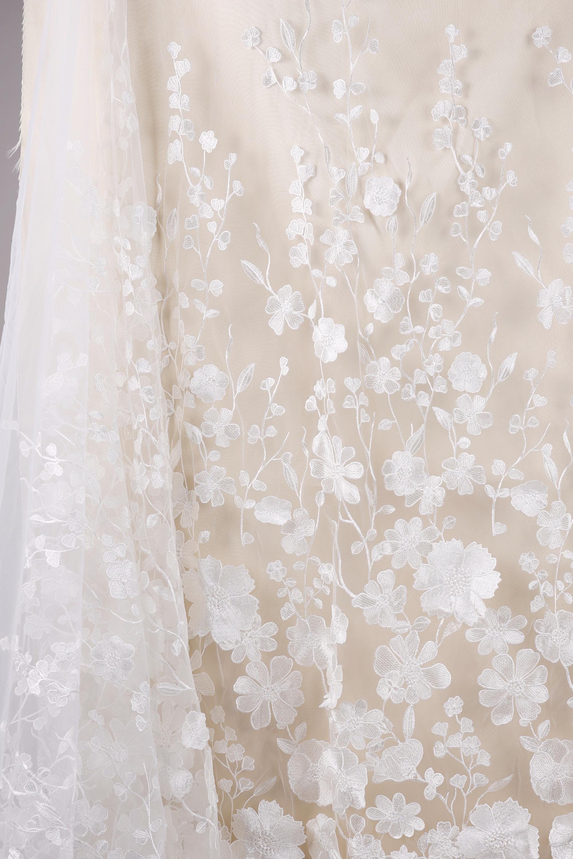 a close-up view of a delicate, white lace fabric with intricate floral embroidery. The fabric appears to be a wedding dress or a similar elegant garment, with the lace and floral patterns creating a visually appealing and delicate design.