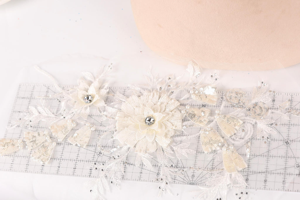 a close-up view of a delicate, white lace flower with a crystal center, surrounded by additional lace flowers and leaves. The flowers are arranged on a transparent surface, which appears to be a ruler or measuring tool.