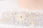 a close-up view of a delicate, white lace flower with a crystal center, surrounded by additional lace flowers and leaves. The flowers are arranged on a transparent surface, which appears to be a ruler or measuring tool.