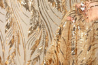 a close-up view of a gold sequin fabric with a floral pattern. The fabric appears to be a sheer material, with a gold and black color scheme. The background is a plain white wall, which contrasts with the vibrant colors of the fabric. The image does not contain any text or other discernible objects.