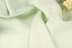 A close-up view of a soft, light green fabric, possibly linen or chiffon, with a delicate flower visible in the background.