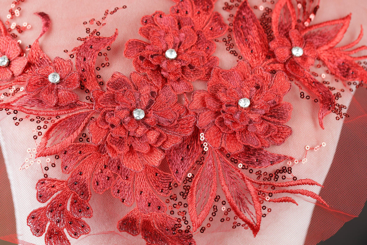 a close-up view of a delicate, intricately embroidered floral design on a fabric. The design is composed of numerous red flowers, each adorned with a small crystal or gemstone, creating a striking and eye-catching visual effect. The flowers are meticulously arranged, with some overlapping each other to create a sense of depth and complexity. The overall composition of the image is visually appealing and showcases the skill and artistry involved in the creation of this embroidered design.