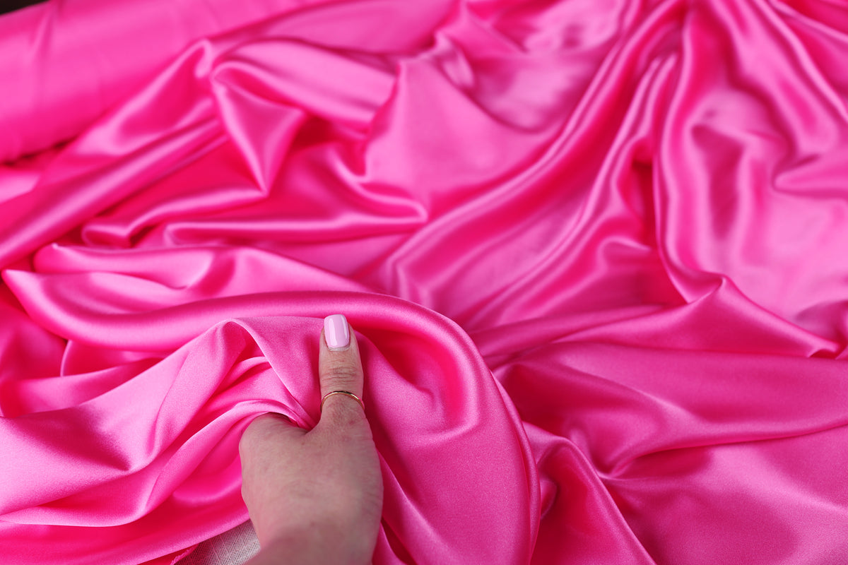 A hand is holding a piece of bright pink satin fabric, which appears to be soft and smooth.