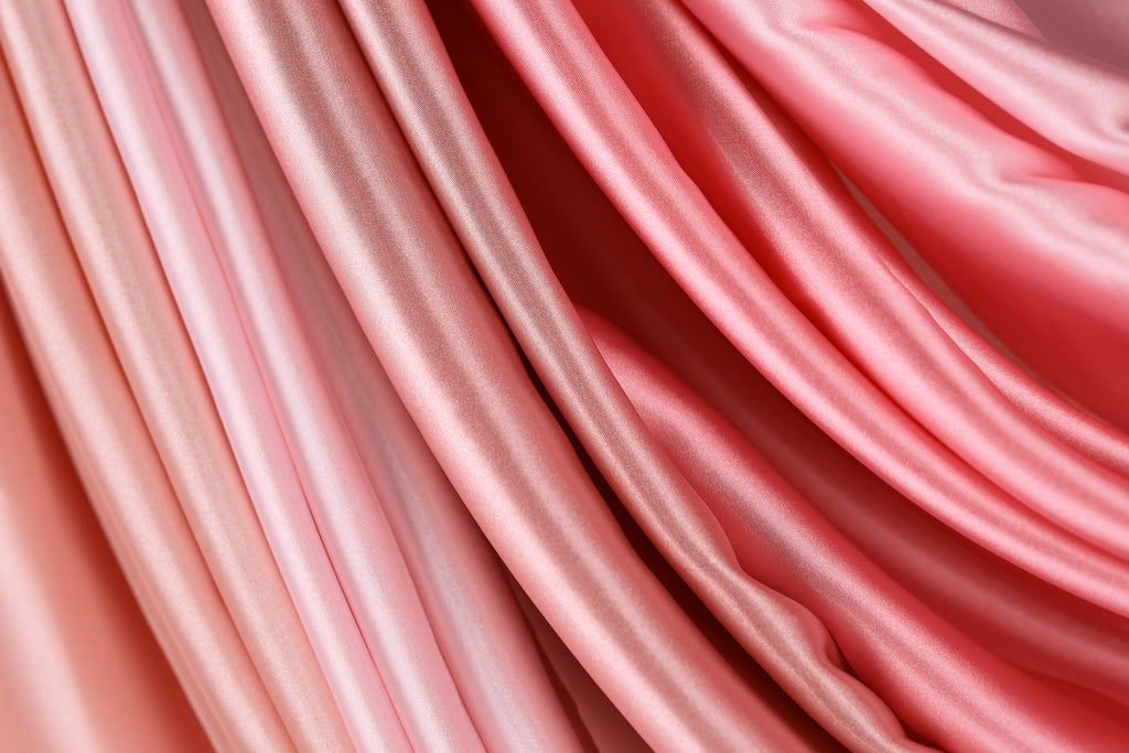 a close-up view of a soft, flowing fabric with a gradient of pink and red colors. The fabric appears to be made of satin or a similar material, with a smooth and shiny texture. The colors are vibrant and the fabric appears to be in good condition, suggesting it is a high-quality material.