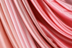 a close-up view of a soft, flowing fabric with a gradient of pink and red colors. The fabric appears to be made of satin or a similar material, with a smooth and shiny texture. The colors are vibrant and the fabric appears to be in good condition, suggesting it is a high-quality material.