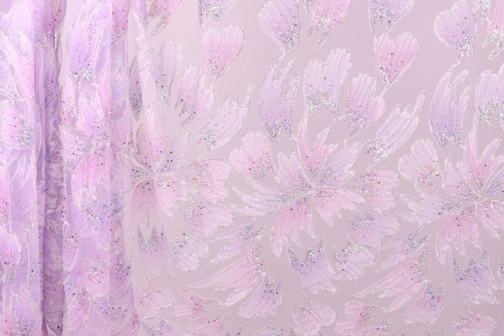 a close-up view of a fabric with a floral pattern in shades of pink and purple. The fabric appears to be made of a sheer material, with a delicate and intricate design. The pattern consists of numerous small flowers or petals, each with a unique shape and color, creating a visually appealing and textured appearance. The fabric is displayed against a light pink background, which further accentuates the vibrant colors and intricate details of the floral pattern.