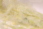 a close-up view of a delicate, sheer fabric with a yellow floral pattern. The fabric appears to be made of sheer material, giving it a soft and delicate appearance. The pattern consists of small yellow flowers or leaves, which are intricately designed and embellished with small, colorful dots or sequins. The fabric is spread out, revealing its intricate details and textures, creating a visually appealing and delicate piece of textile art.