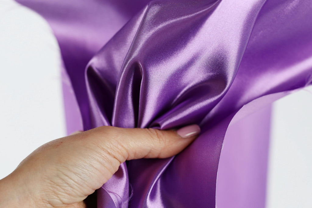 A hand is holding a piece of purple satin fabric, with the fabric being folded and twisted in a way that it appears to be being held or manipulated.