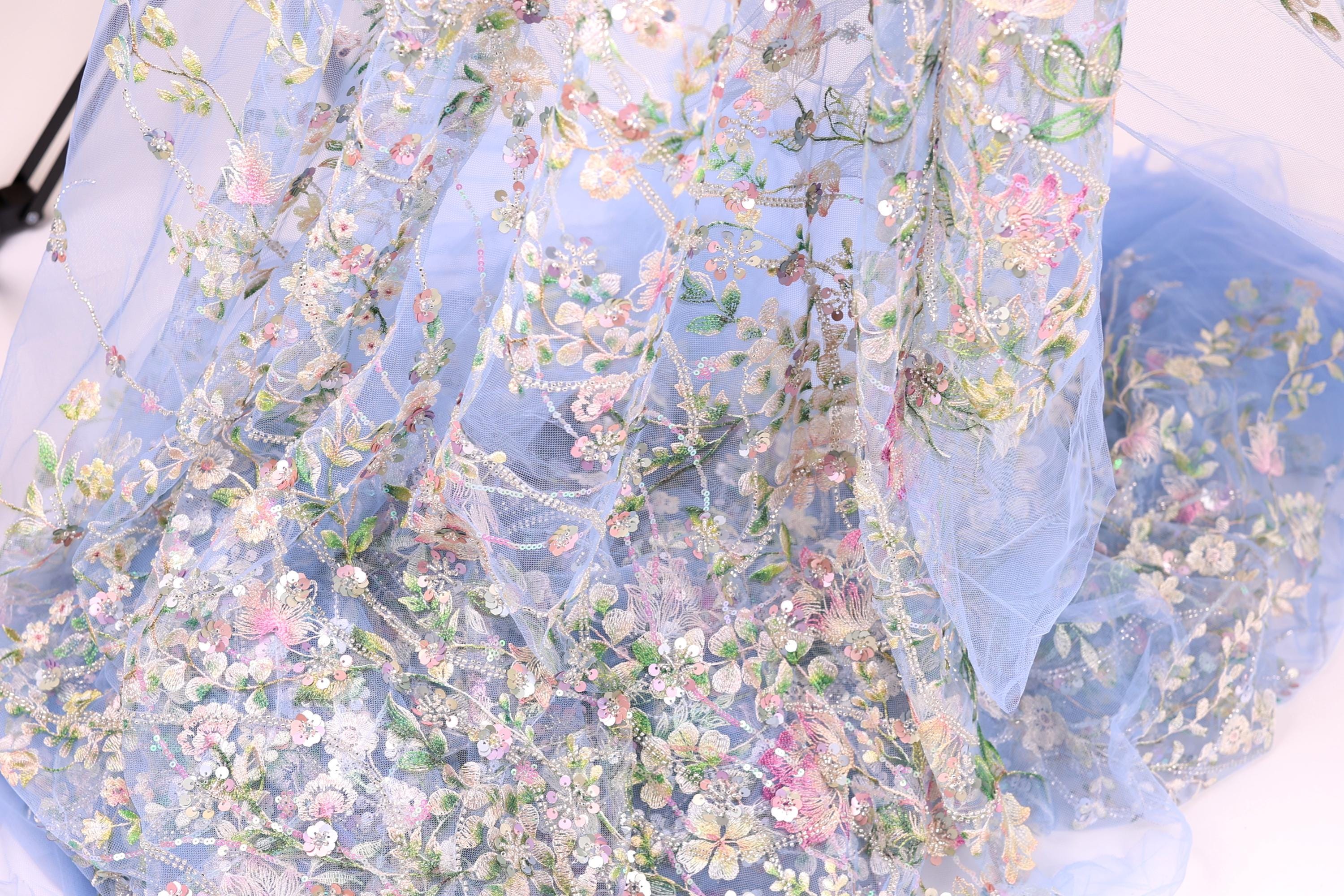 a close-up view of a delicate, intricately embroidered fabric with a floral pattern. The fabric appears to be a light blue color, and the embroidery features a variety of flowers and leaves in shades of pink, white, and green. The fabric is intricately detailed, with the flowers and leaves appearing to be hand-stitched, creating a visually appealing and delicate appearance.