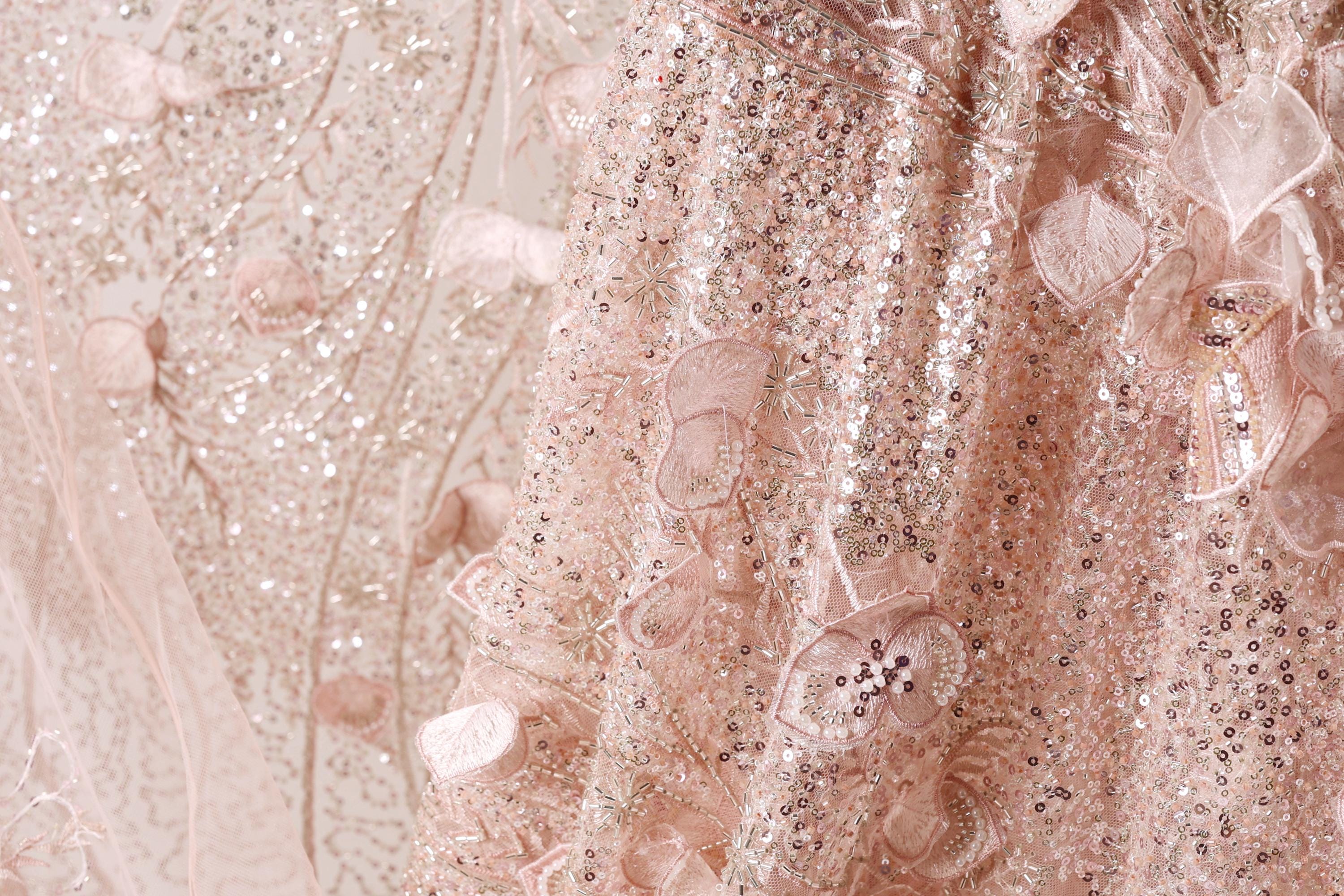 The image is a close-up of a delicate, pink, and beige fabric with intricate details, including sequins and lace-like patterns.