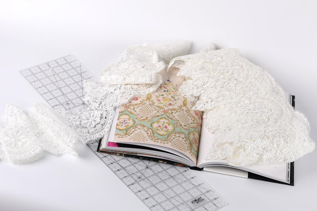 an open book with a lace pattern on the cover, lying on a white surface. There is also a piece of white lace and a ruler visible in the scene.