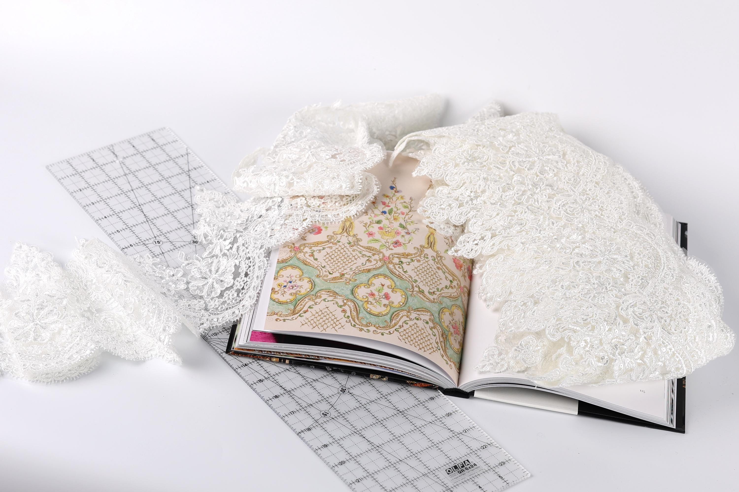 an open book with a lace pattern on the cover, lying on a white surface. There is also a piece of white lace and a ruler visible in the scene.