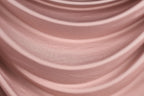 a close-up view of a soft, flowing pink fabric, possibly a satin or chiffon material. The fabric has a smooth, wavy texture that creates a sense of movement and softness.