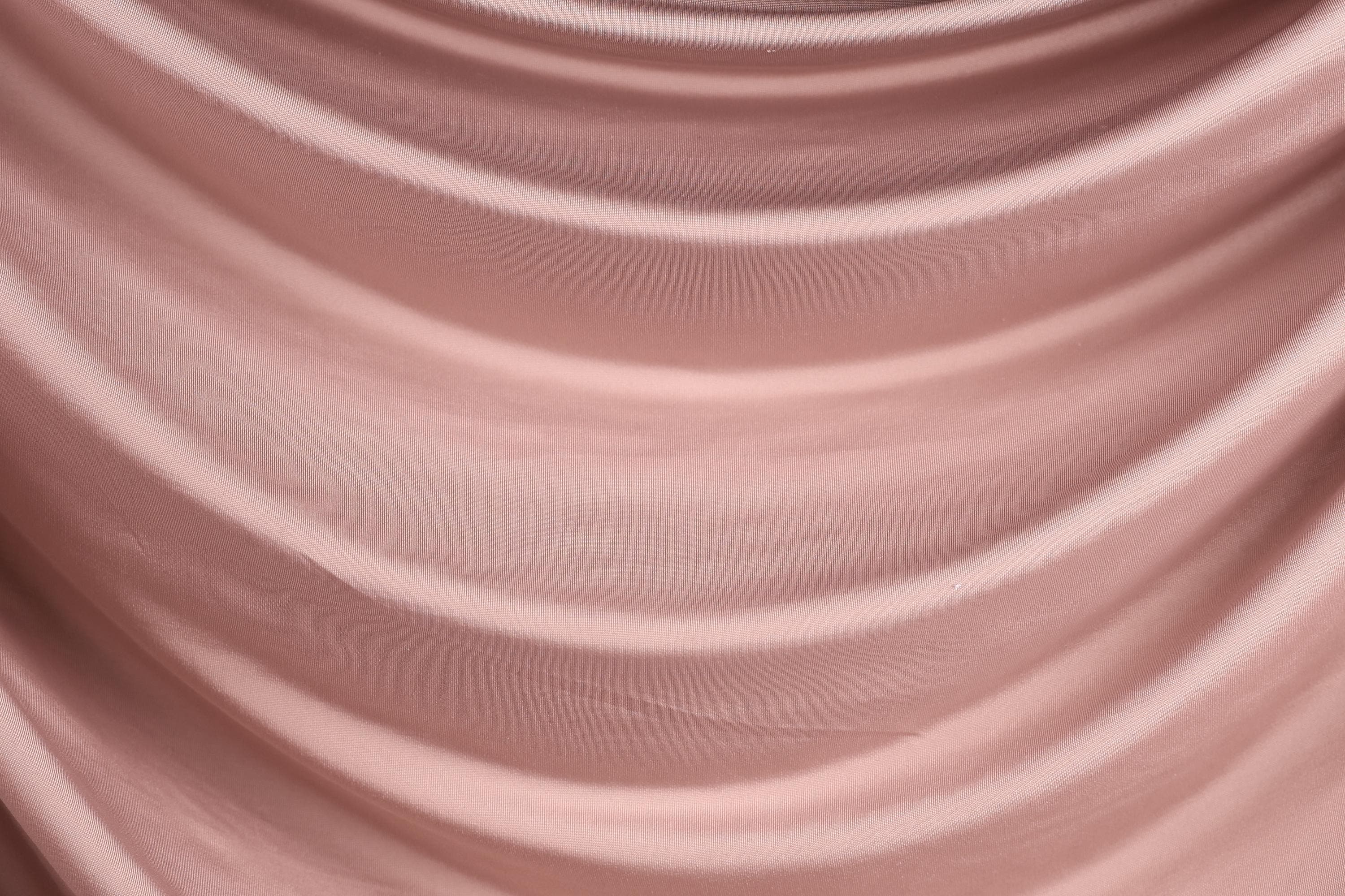 a close-up view of a soft, flowing pink fabric, possibly a satin or chiffon material. The fabric has a smooth, wavy texture that creates a sense of movement and softness.