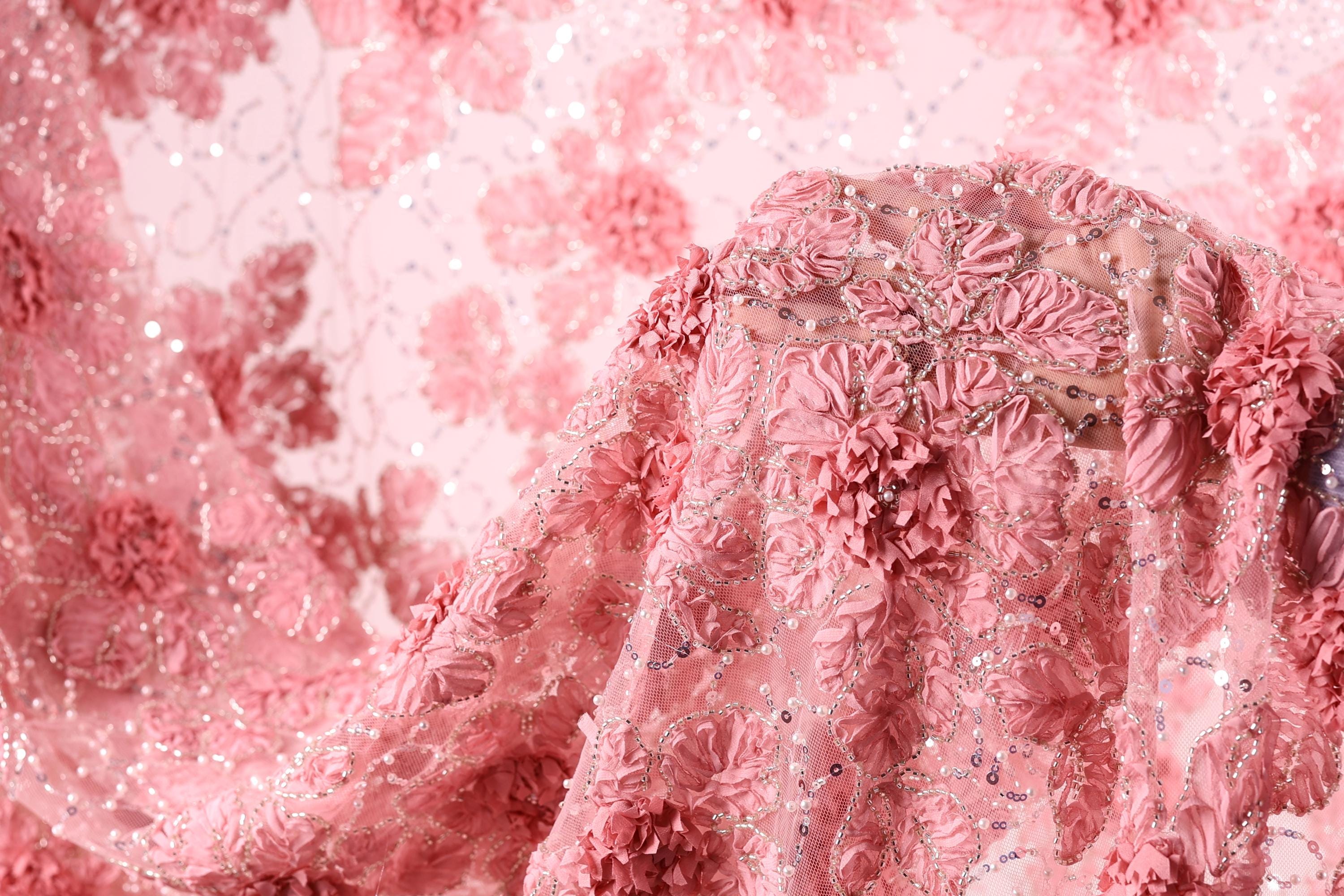 a close-up view of a delicate, pink fabric with intricate floral designs. The fabric appears to be made of lace or a similar delicate material, with a floral pattern that includes pink flowers and leaves. The fabric is draped over a surface, creating a visually appealing and elegant display.