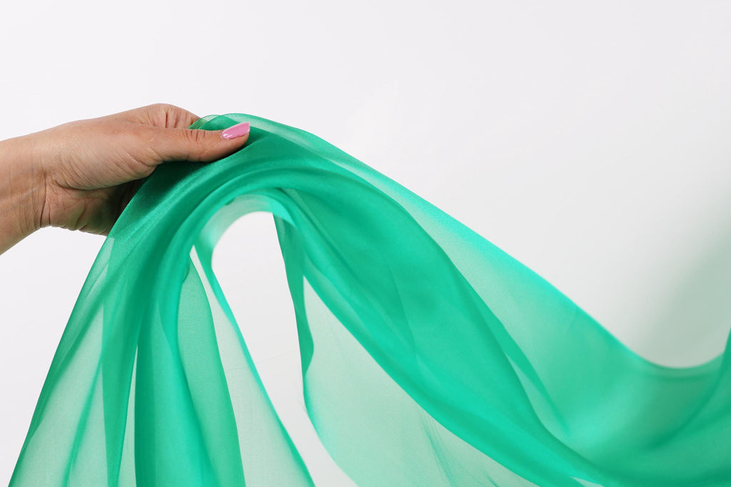 A hand is holding a green fabric, possibly silk or chiffon, against a white background.