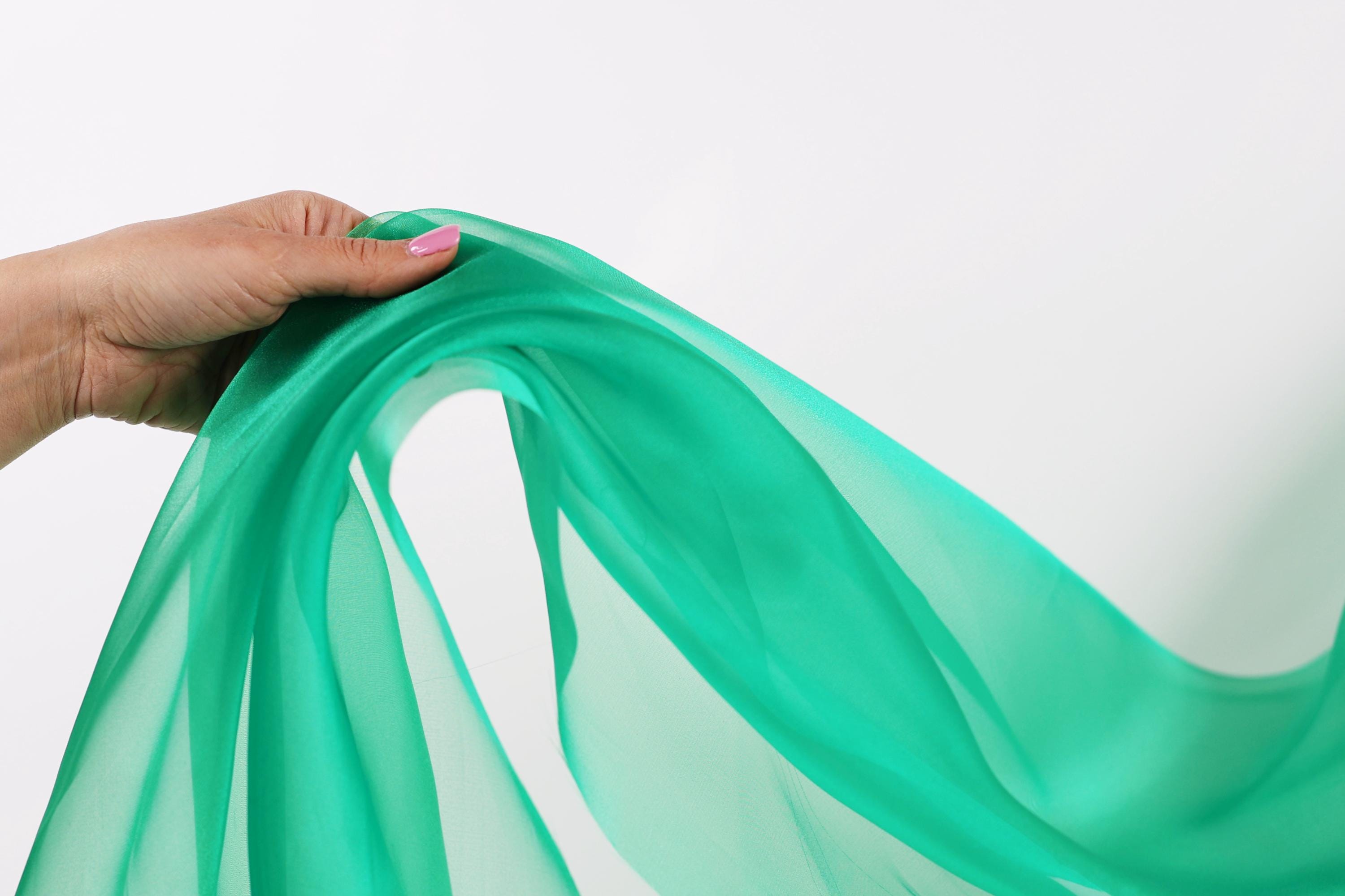 A hand is holding a green fabric, possibly silk or chiffon, against a white background.