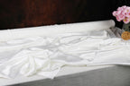 a white fabric, possibly silk or satin, lying on a surface, with a vase of pink flowers placed next to it.